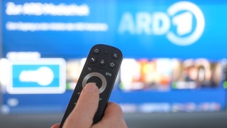 Eine Fernbedienung, dahinter verschwommen ein Fernseher mit dem ARD-Programm