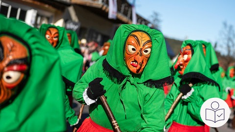Verkleidete Hexen mit grünem Umhang. Die Hexe hat eine Holz·maske. Die Hexe hat einen Besen in der Hand. Rechts das SWR Logo für Leichte Sprache.