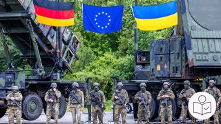 Deutsche und ukrainische Soldaten bei einer Ausbildungs·mission am Flug·abwehr·system "Patriot" auf einem deutschen Truppen·übungs·platz. Über ihnen die Flaggen von der EU, Deutschland und der Ukraine. Rechts das SWR Logo für Leichte Sprache.    