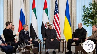 Das ist ein Beispiel·bild: Mehrere Männer sitzen von den Flaggen von der Ukraine, Russland und den USA. Sie reden miteinander. 