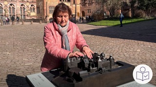 Eine Frau tastet an einem Tast·modell. Das Tast·modell ist vom Schloss Heidelberg. Das Tast·modell ist neu. Das Modell ist aus Metall. Rechts das SWR Logo für Leichte Sprache.