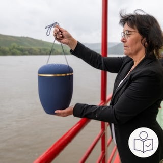 Eine Frau hält eine Urne in den Händen. Die Frau steht am Rand von einem Schiff. im Hintergrund sieht man die Mosel und Weinberge. Rechts das SWR Logo für Leichte Sprache.  