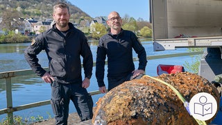 Das Bild ist auf einem Schiff aufgenommen. Vorne liegt eine große Bombe. Sie ist sehr dreckig. Zwei Fach·leute stehen hinter der Bombe. Im Hintergrund sieht man Wasser und Ufer. Rechts das SWR Logo für Leichte Sprache.  
