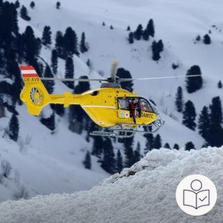 Das ist ein Beispiel·bild: Ein gelber Rettungs·hubschrauber fliegt in den Bergen. Auf den Bergen liegt Schnee. Die Bergrettung hat einen Mann aus Ettlingen aus den Bergen gerettet. Rechts das SWR Logo für Leichte Sprache.