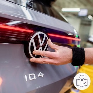 Ein VW-Mitarbeiter montiert das Firmenlogo auf der Rückseite eines Elektroautos. Rechts das SWR Logo für Leichte Sprache.