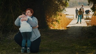 Ein Kind und eine Frau umarmen sich, im Hintergrund angeln zwei Männer und zwei Jungen auf einem Steeg an einem See