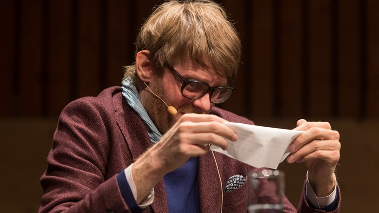 Mann auf Podium gestikuliert mit Stück Papier