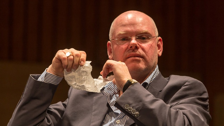 Mann mit Brille schaut frontal in die Kamera und hält ein zerknittertes Papier