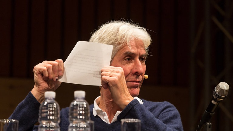 Mann auf Podium sitzend mit Mikroport hält Blatt Papier hoch