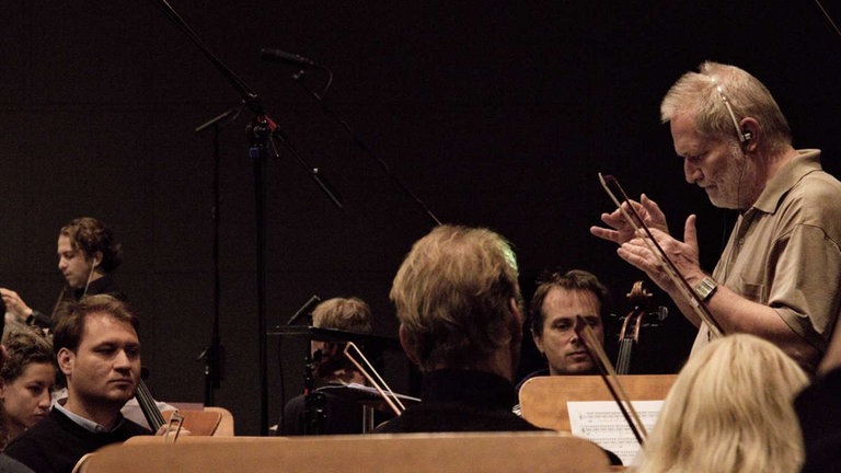 Mann mit Bart und mit Headset dirigiert Orchester