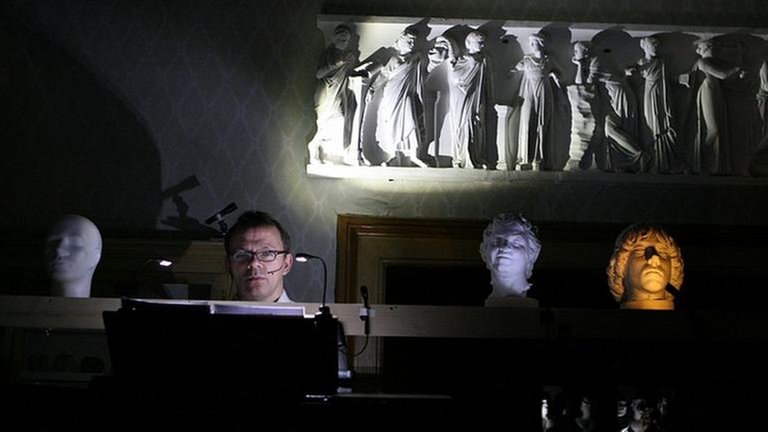 Sprecher sitzt hinter einem Pult in einem angedeuteten Wohnzimmer, neben ihm stehen mehrere antike Gipsköpfe