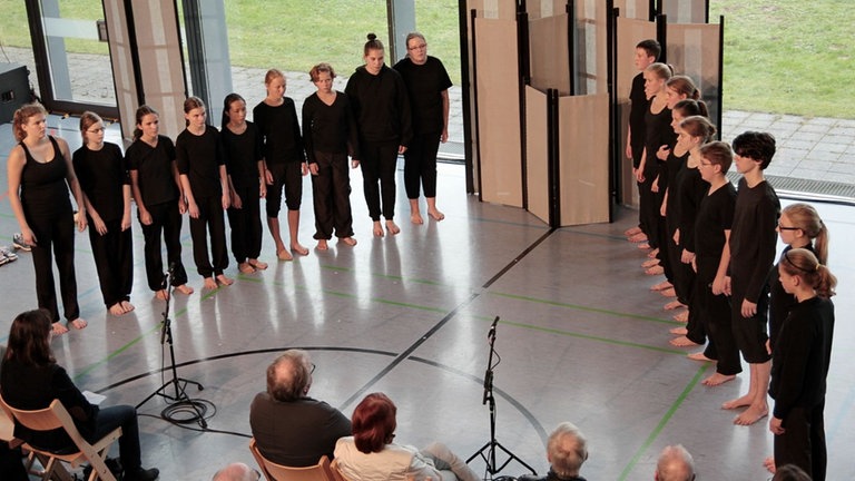 Schwarz gekleidete Schüler*innen stehen sich in zwei spitz zulaufenden Reihen 