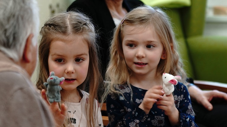 Zwei Mädchen aus dem Kindergarten zeigen einer Seniorin ihre Fingerpuppen.