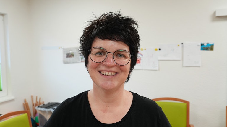 Frau mit kurzen, dunklen Haaren und Brille in einem Raum mit Stühlen.