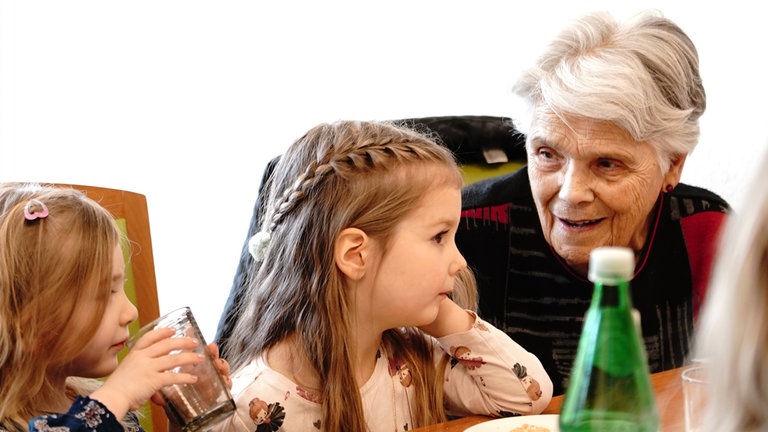 Eine ältere Frau mit kurzen grauen Haaren und schwarz-roten Pullover redet mit zwei Kindergartenkindern, die neben ihr sitzen.