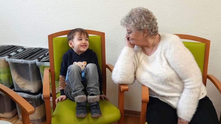 Eine alte Dame in weißem Pullover schaut einem kleinen Jungen zu, wie er kichernd auf seinem Stuhl sitzt.
