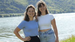 Zwei Frauen mit braunen Haaren und T-Shirt an, umarmen sich und stehen vor einem Fluss und grünen Hängen.