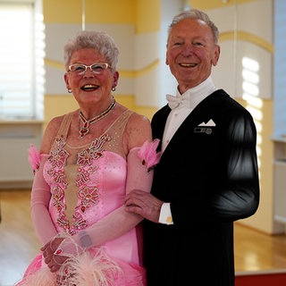 Christa und Karl-Heinz stehen Arm in Arm im Tanzsaal. Christa trägt ein langes, pinkes Tanzkleid. Karl-Heinz einen schwarzen Anzug. 
