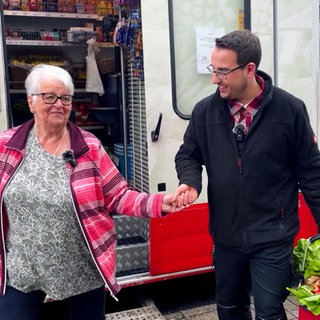 Aaron hilft einer älteren Kundin aus dem Supermarkt-Lastwagen. Er nimmt sie an der Hand und lächelt, während er in der anderen Hand ihren Einkaufskorb mit viel Gemüse trägt. Hinter ihnen sieht man den offenen Supermarkt-Wagen, die Kundin trägt eine rosa-gestreifte Jacke.