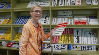 Ältere Dame, mit bunter Brill, steht vor einem Regal, in dem ganz viele Schilder liegen. In ihrer Hand hält sie ein Schild.
