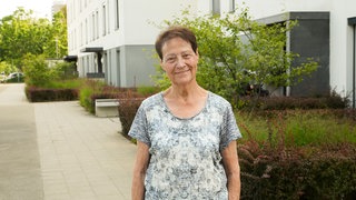 Eine ältere Frau mit braunen Haaren lächelt in die Kamera. Hinter ihr ist ein weißer Wohnkomplex mit vielen Bäumen rundherum zu sehen.