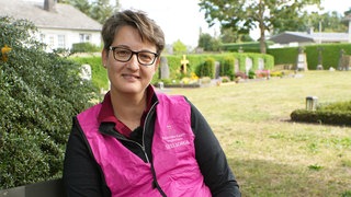 Mittelalte Frau mit braunen Haaren. Sie trägt eine pinke Warnweste.
