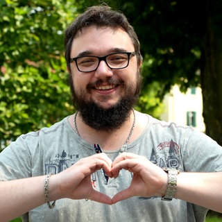 Mann mit Brille lacht in die Kamera und formt ein Herz mit seinen Händen.