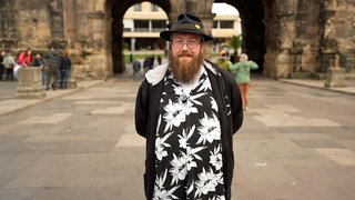 Mann mit Hut und Brille lächelt vor der Porta Nigra in Trier.