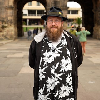 Mann mit Hut und Brille lächelt vor der Porta Nigra in Trier.