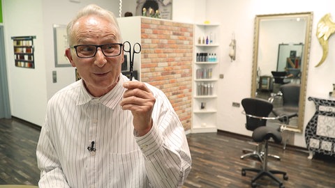 Mann mit Brille und Schere in der Hand lacht im Friseurladen.