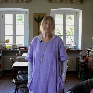 Eine Frau mit blonden Haaren und lila Kleid lächelt in die Kamera.