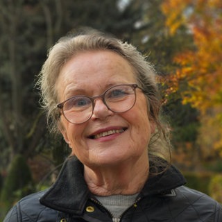 Ältere Dame mit grauen langen Haaren und brille lächelt an einem Herbsttag in die Kamera.