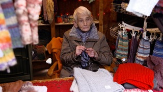 Ältere Dame sitzt in einem Weihnachtsmarkt Häuschen und verkauft Stricksachen.