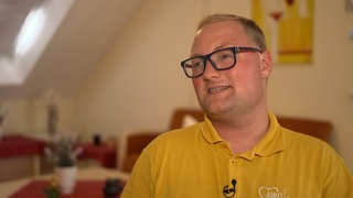 Junger Mann mit blonden Haaren, Brille und gelbem T-Shirt lächelt.