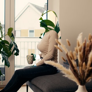 Katrin sitzt mit einer Tasse in der Hand auf einem Sofa. Sie schaut geradeaus. Ihr Gesicht und ihre Hände sind unkenntlich gemacht.