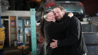 Isabella und Chris sind seit 14 Jahren verheiratet und haben einen Altersunterschied von 16 Jahren. Die beiden stehen in der Werkstatt ihres Abschleppunternehmens. Im Hintergrund ist ein Auto auf einer Hebebühne zu sehen. Isabella und Chris stehen davor und umarmen sich. Beide lächeln glücklich in die Kamera.