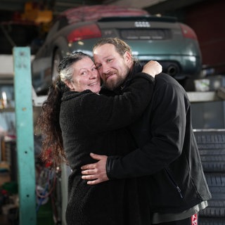 Isabella und Chris sind seit 14 Jahren verheiratet und haben einen Altersunterschied von 16 Jahren. Die beiden stehen in der Werkstatt ihres Abschleppunternehmens. Im Hintergrund ist ein Auto auf einer Hebebühne zu sehen. Isabella und Chris stehen davor und umarmen sich. Beide lächeln glücklich in die Kamera.