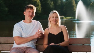 Das Paar Isabel und Aaron haben einen Campingplatz in Isny übernommen. Sie sitzen auf einer Bank beim Felderholweiher und schauen in die Kamera.