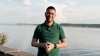 Taha steht vor dem Bodensee und strahlt in die Kamera. 