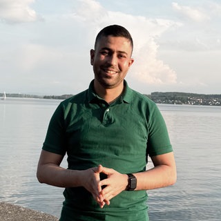 Taha steht vor dem Bodensee und strahlt in die Kamera. 