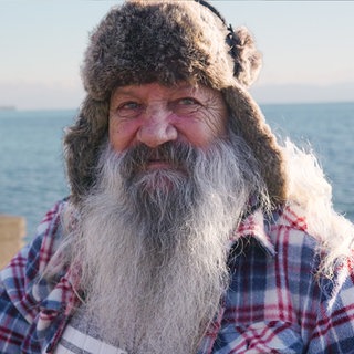 Manne, ehemalig obdachlos, sitzt im Rollstuhl vor dem Bodensee. Mit seinem langen braunen Bart blickt er direkt in die Kamera.