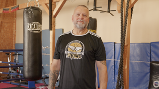 Boxtrainer Harald steht in einem Boxclub. Er trägt Sportkleidung und hat einen grauen Bart. Er schaut direkt in die Kamera. 
