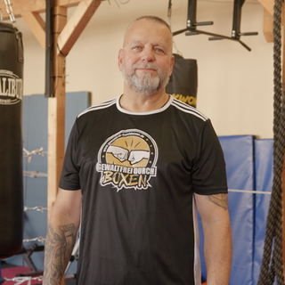 Boxtrainer Harald steht in einem Boxclub. Er trägt Sportkleidung und hat einen grauen Bart. Er schaut direkt in die Kamera. 