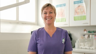 Natalie arbeitet seit 13 Jahren als Pflegerin in der JVA. Natalie steht in einem Behandlungszimmer und trägt einen blauen Kasack. Sie hat kurze blonde Haare und schaut freundlich in die Kamera.