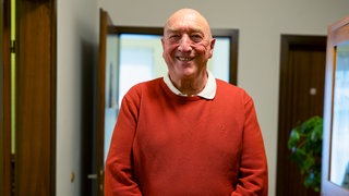 Franz steht in der Teestube in Friedrichshafen, bei der er ehrenamtlich arbeitet. Er trägt einen roten Pullover und lacht in die Kamera.