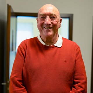 Franz steht in der Teestube in Friedrichshafen, bei der er ehrenamtlich arbeitet. Er trägt einen roten Pullover und lacht in die Kamera.