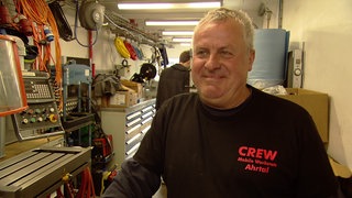 Älterer Mann, mit schwarzem Pulli steht in einer länglichen Werkstatt. Im Hintergrund Maschinen