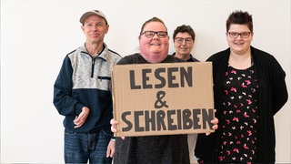 Vier Menschen einer Selbsthilfegruppe lächelnd vor einer weißen Wand