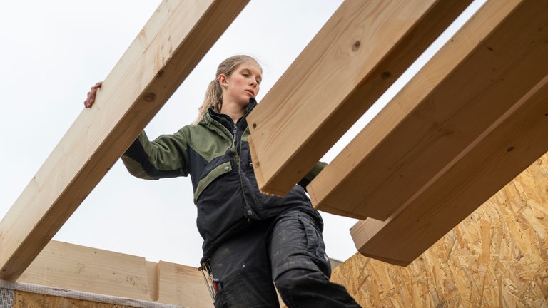 Eine junge, blonde Frau steht vor mehreren Holzbalken, aus denen eine Decke gebaut wird.