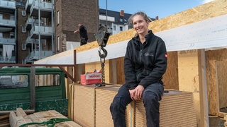 Eine junge, blonde Frau sitzt lächelnd in Arbeitskleidung auf einem Stapel OSB-Platten auf einer Baustelle.
